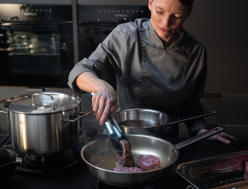 A chef in gray uses tongs to sear steaks in a Fissler frying pan with Fissler stainless steel pot and pan in an elegant modern kitchen.