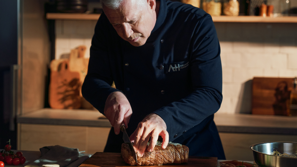 A chef in a dark uniform expertly cuts bread on a wooden board, Fissler cookware and utensils are visible in the warmly lit kitchen.