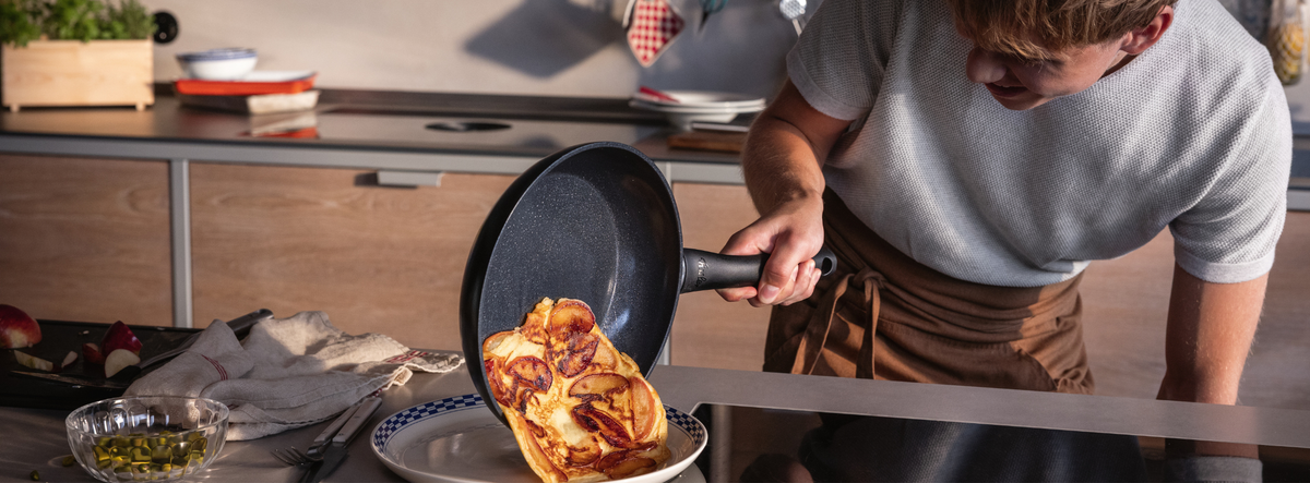 Eine Person gleitet fachmännisch ein goldenes Omelett aus einer Fissler-Pfanne auf einen Teller; hochwertiges Kochgeschirr und Utensilien stehen auf der Arbeitsplatte.