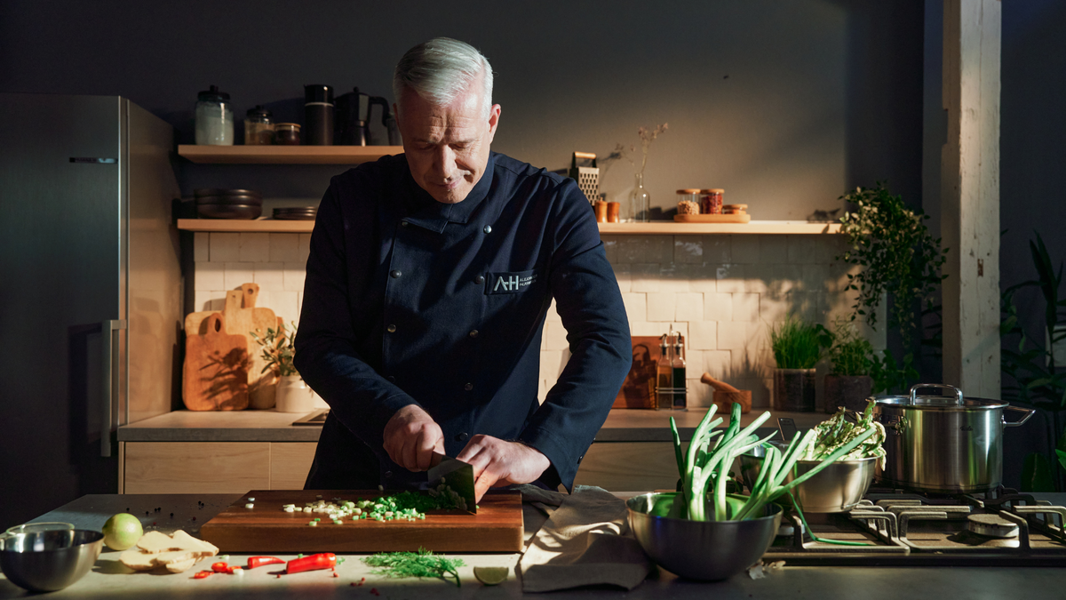 A chef in a dark uniform cuts green onions on a wooden board, next to it are stainless steel cookware from Fissler and fresh vegetables.