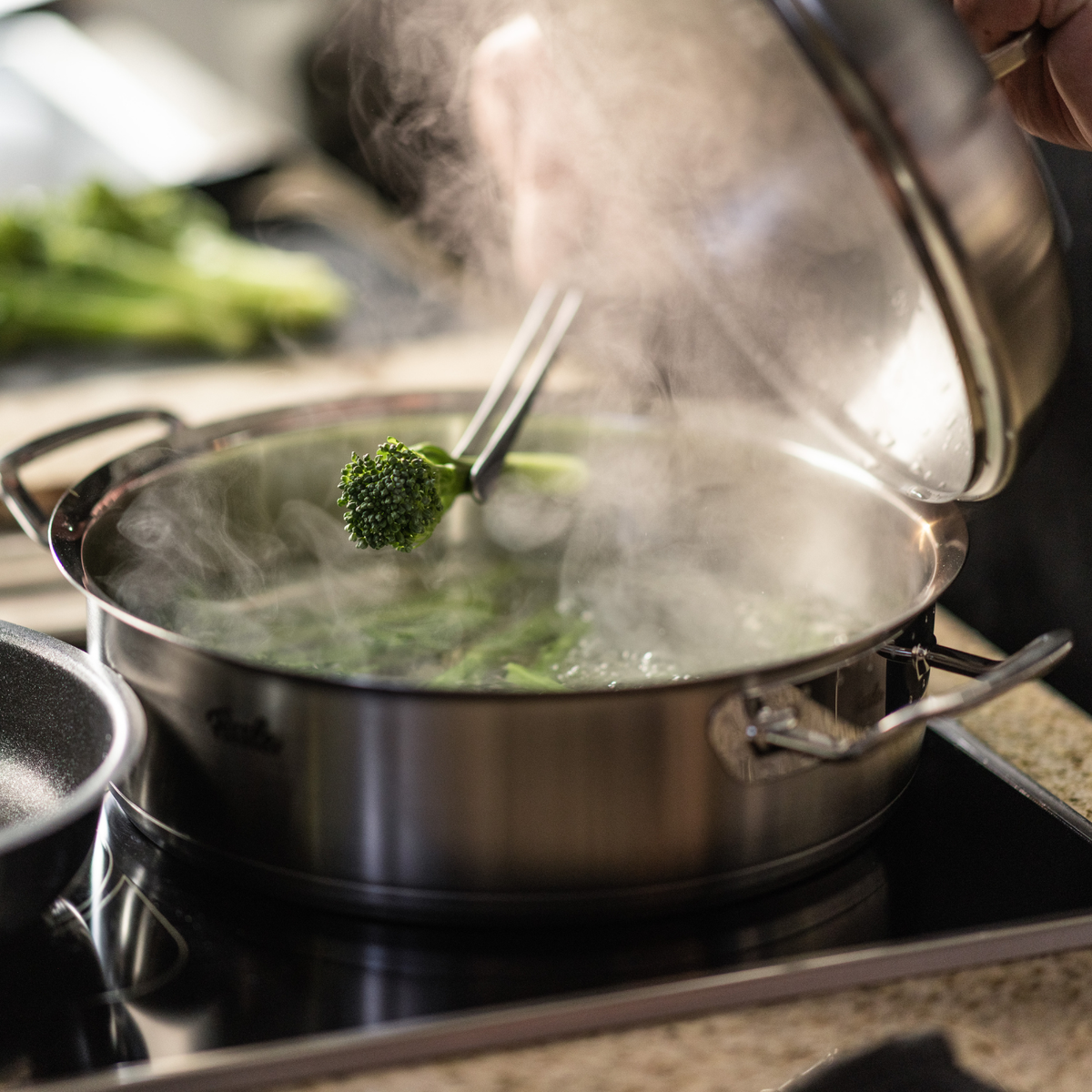 Ein Koch öffnet einen Fissler-Edelstahltopf, der auf dem Herd dampft, und hält mit einer Zange Brokkoli fest. Frisches Gemüse unterstreicht die Qualität der Küche.