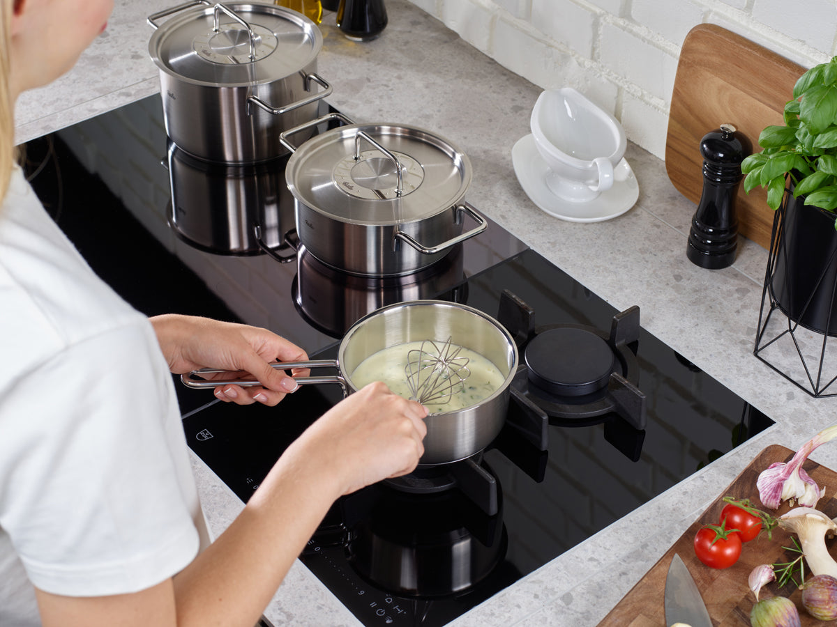 Eine Person rührt eine cremige Soße in einer Fissler Pure Collection Stielkasserolle aus hochwertigem Edelstahl auf einem modernen Herd um, umgeben von weiteren Fissler-Töpfen, frischen Kräutern und Gemüse auf der Küchenarbeitsplatte. Diese Szene unterstreicht Fisslers renommierte deutsche Ingenieurskunst, das zeitlose Design und die hervorragende Qualität des Kochgeschirrs – eine Tradition, auf die man seit 1845 vertraut.