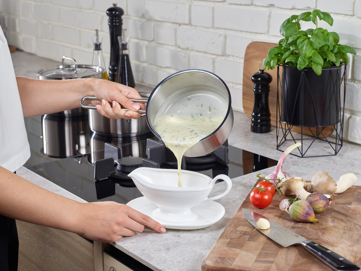 In einer modernen Küche gießt eine Person elegant eine cremige Kräutersauce aus einer Fissler Pure Collection Stielkasserolle – ein Beispiel für präzise Technik und zeitloses Design „Made in Germany“ – in eine Sauciere. Auf der Arbeitsplatte unterstreichen frisches Gemüse, Basilikum und ein Schneidebrett die einladende Atmosphäre, die durch das hochwertige Edelstahl-Kochgeschirr von Fissler entsteht – bekannt für Langlebigkeit und Innovation seit 1845.