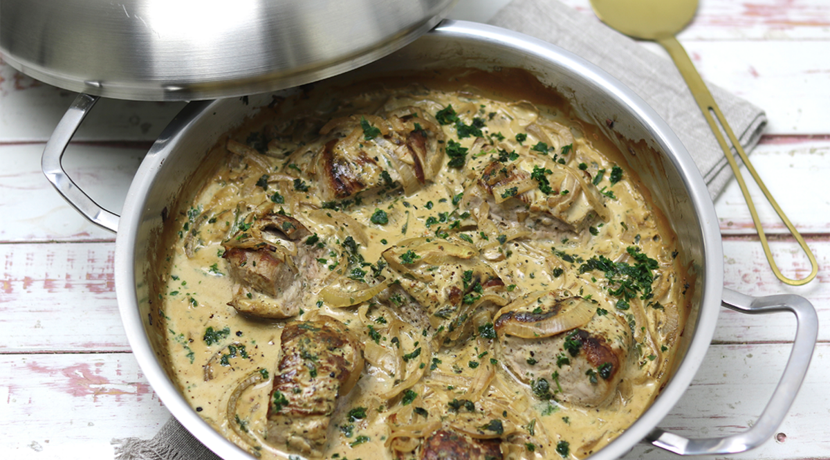 Fissler-Edelstahlpfanne mit Schweinemedaillons in Sahnesauce, Kräutern und Zwiebeln; teilweise mit Deckel bedeckt. Goldlöffel in der Nähe.