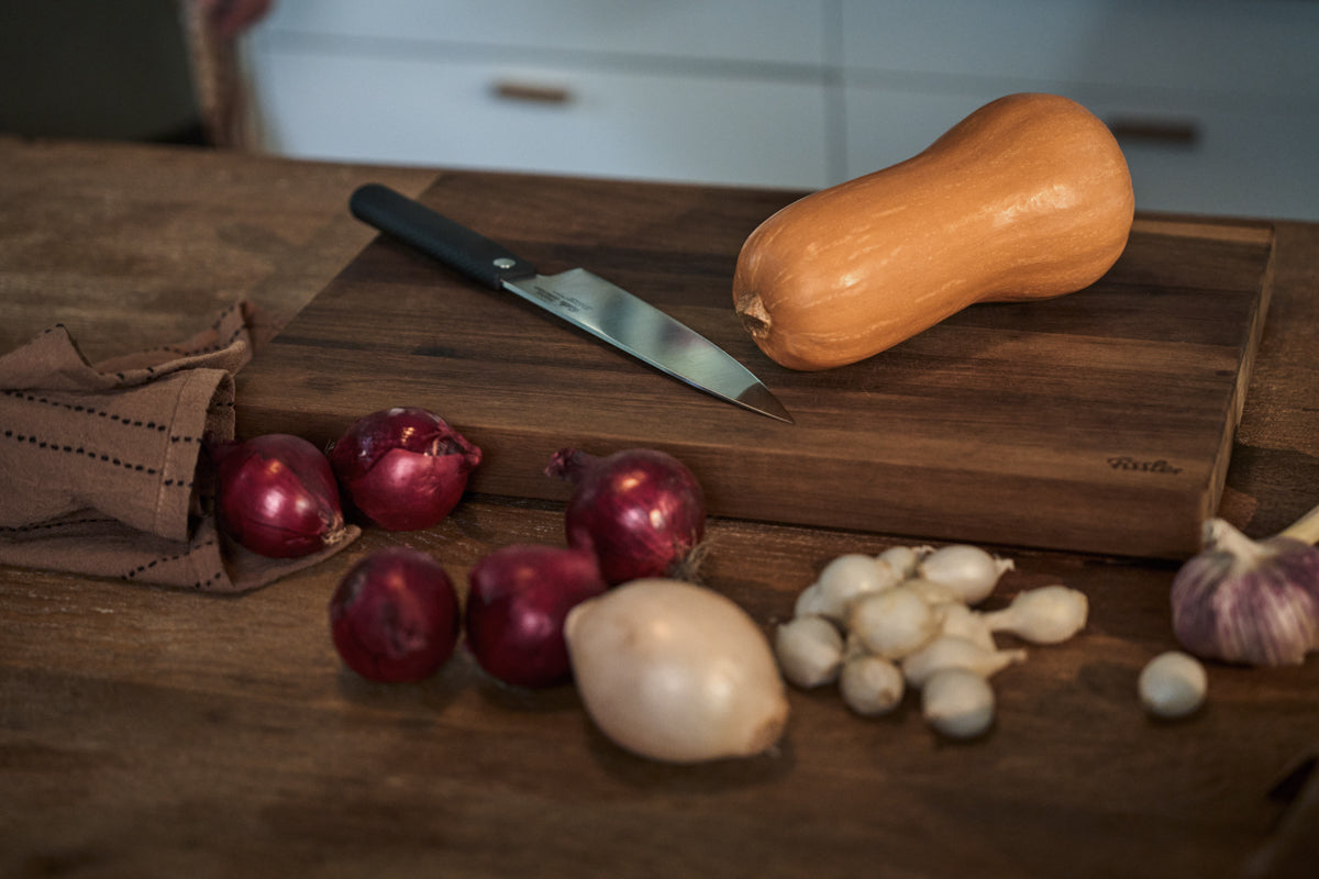 A butternut squash, onions, garlic, and a premium knife are arranged on a wooden Fissler cutting board atop a table. A neatly folded cloth rests nearby, all set within an inviting kitchen—reflecting the precision, durability, and timeless design of Fissler quality kitchenware.