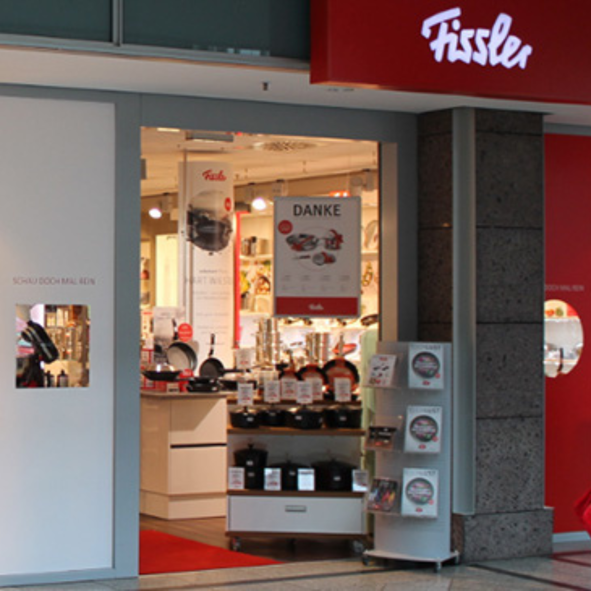 Eingang zu einem Fissler-Geschäft, in dem hochwertiges deutsches Kochgeschirr präsentiert wird, "DANKE"-Schild, Broschüren und Produktauslagen aus Edelstahl.