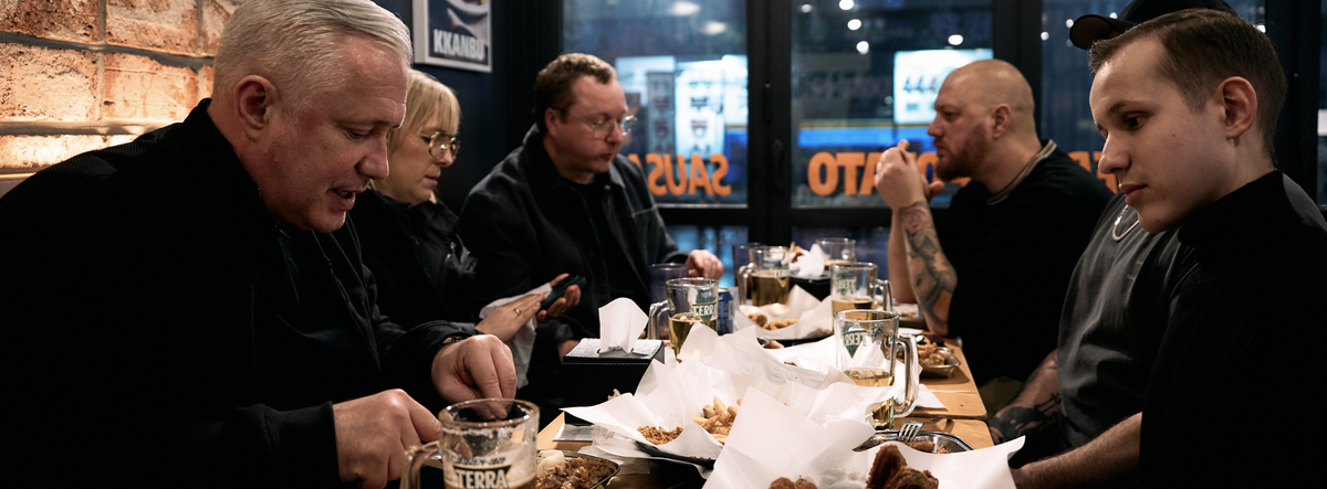 Eine Gruppe von Menschen versammelt sich in einem gemütlichen Restaurant um einen Holztisch und unterhält sich bei fröhlichem Gelächter. Die erlesenen Speisen sind zusammen mit Getränken und Gewürzen wunderschön auf den Tellern angerichtet, wobei Fisslers meisterhaft gefertigtes Edelstahl-Kochgeschirr – wie Töpfe und Pfannen – dezent in die Tischdekoration integriert ist. Das Ambiente ist warm und einladend, unterstrichen durch sanfte Beleuchtung und große Fenster, und spiegelt Fisslers Engagement für