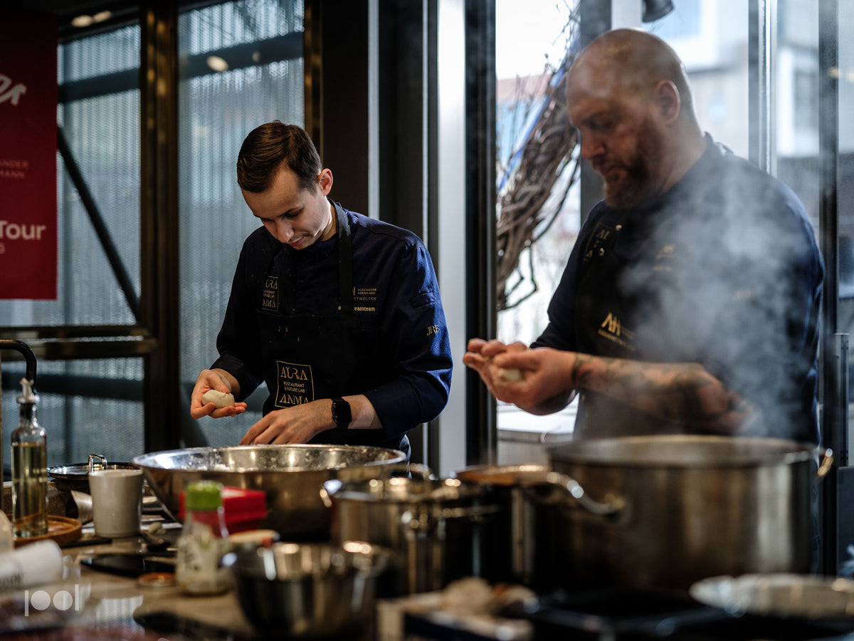 In einer hellen, von Tageslicht durchfluteten Profiküche bereiten zwei Köche in dunklen Uniformen Speisen mit hochwertigem Fissler-Edelstahl-Kochgeschirr zu. Ein Koch formt gekonnt Teig neben glänzenden Fissler-Töpfen und -Pfannen, während der andere sich um die dampfenden Schnellkochtöpfe auf dem Herd kümmert. Auf der Arbeitsfläche sind eine Auswahl an langlebigen Fissler-Küchenutensilien und frische Zutaten zu sehen, die für präzise Technik und zeitloses deutsches Design stehen.