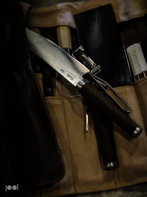 Ein Fissler-Kochmesser mit einem dunklen, ergonomisch geformten Griff liegt auf einer hellbraunen Messerrolle, flankiert von hochwertigen Küchenutensilien – einem Präzisionsschäler, einem zuverlässigen Wetzstahl und einem robusten schwarzen Hackmesser –, die Fisslers Bekenntnis zu deutscher Handwerkskunst und zeitlosem Design verdeutlichen.